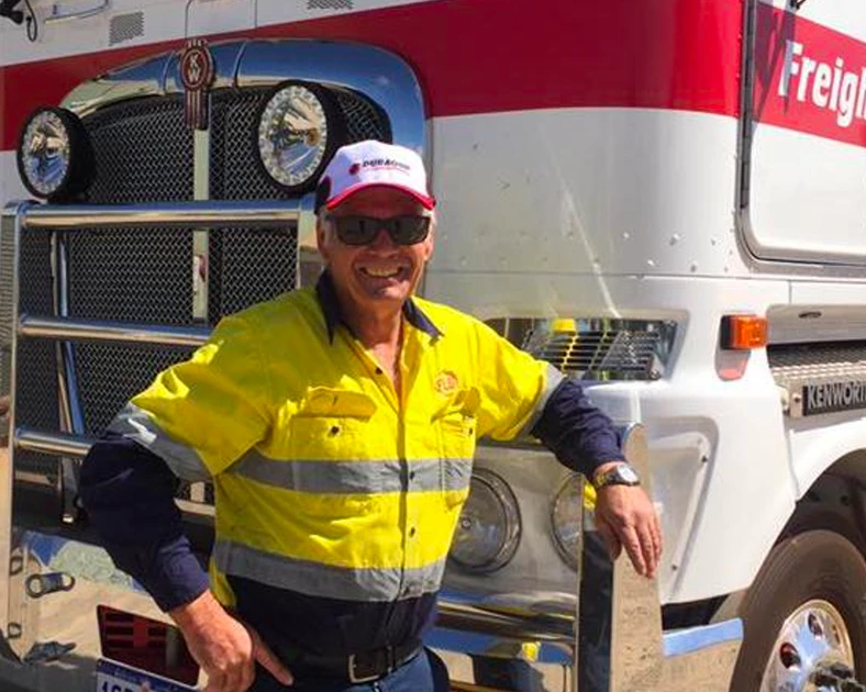 A truck driver wearing high-vis yellow shirt and white cap leaning against the bumper of his large semi truck and trailer.