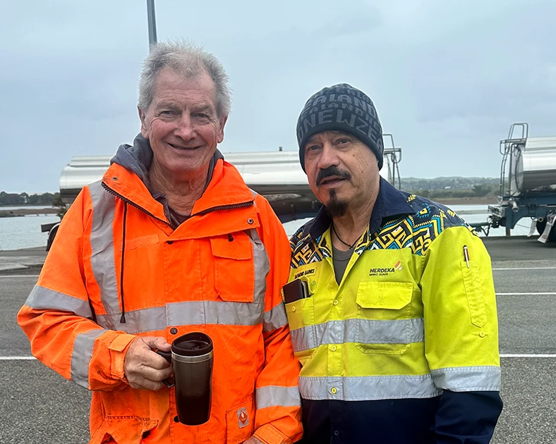 Two truck drivers wearing high-vis clothing smiling at the camera. One is holding a travel mug of coffee. A large truck can be seen in the background.